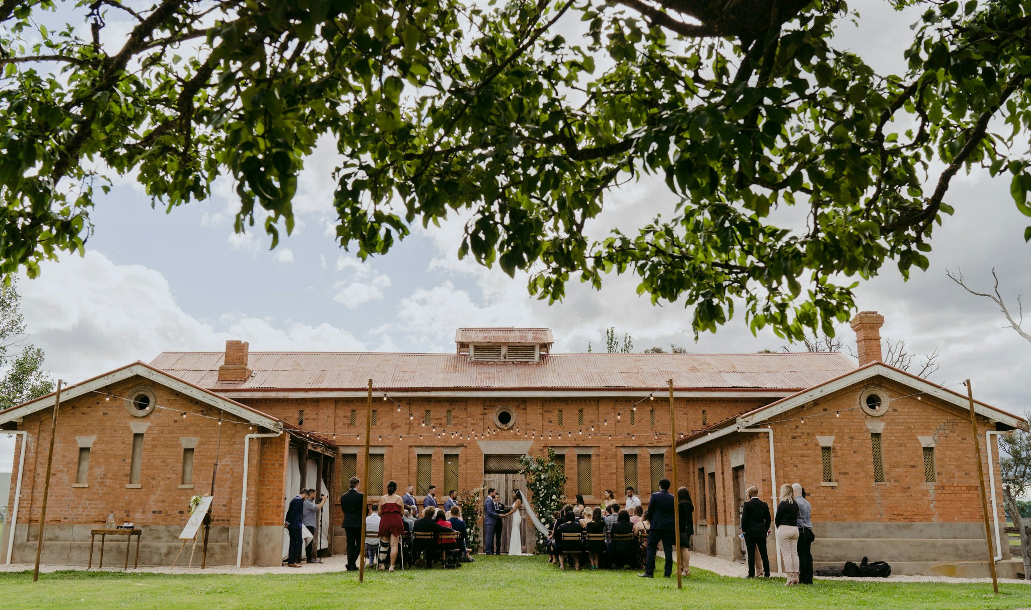 Wedding ceremony underway, in front of brick stables.